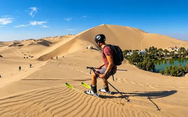 Man wearing a helmet and backpack sand skiing down a high dune with Huacachina oasis and lagoon in the background.