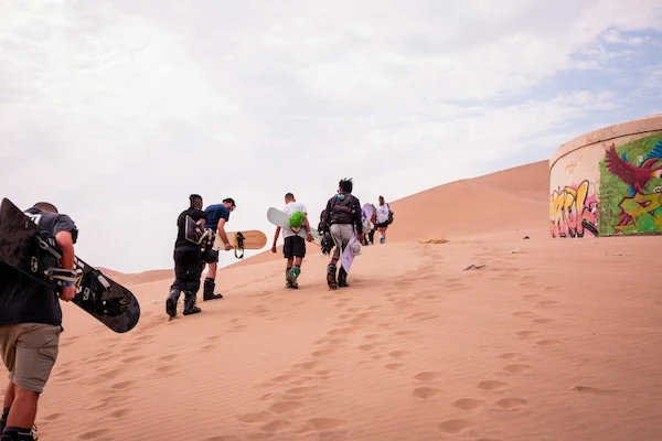 A group of tourists walking through the Huacachina dunes, carrying their sandboards and wearing comfortable sportswear.